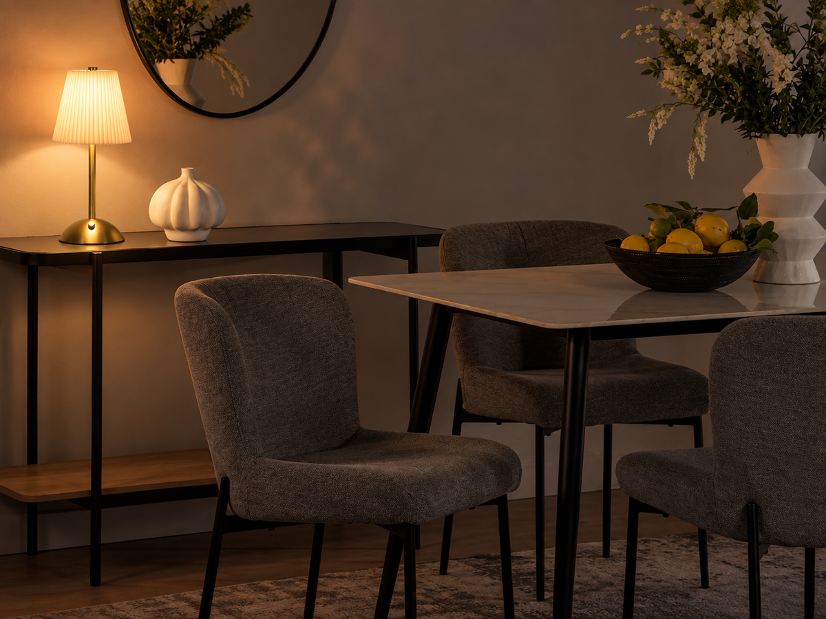 A modern dining room scene with gray upholstered chairs around a light stone dining table, a black console table with a lit lamp and white vase, and a bowl of lemons on the table. The background includes a warm wall, round mirror, and floral arrangement in a white vase.