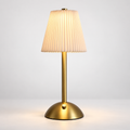 A modern table lamp with a pleated beige fabric shade, a slim gold metal stem, and a round gold base, shown lit against a clean light gray background.