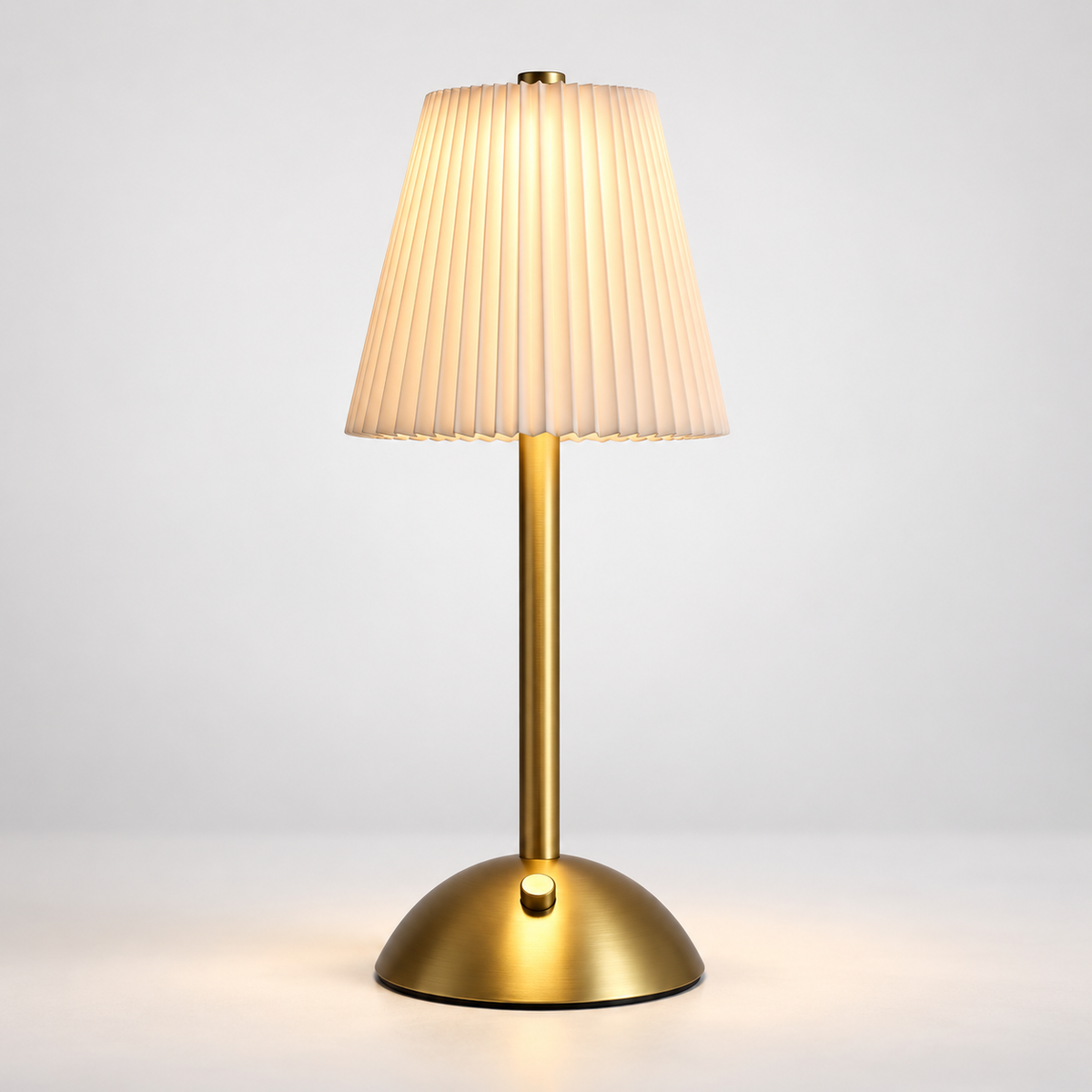 A modern table lamp with a pleated beige fabric shade, a slim gold metal stem, and a round gold base, shown lit against a clean light gray background.