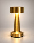 A modern table lamp with a brushed brass finish, featuring a cylindrical shade, a tapered stem, and a circular base, shown illuminated against a neutral gray background.