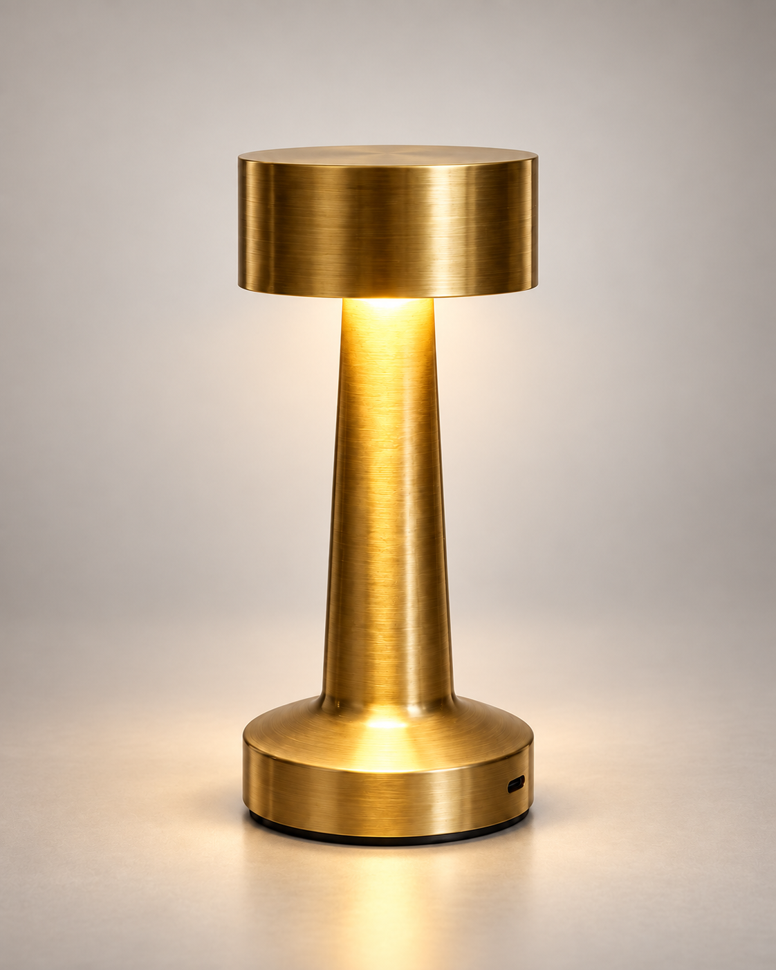 A modern table lamp with a brushed brass finish, featuring a cylindrical shade, a tapered stem, and a circular base, shown illuminated against a neutral gray background.