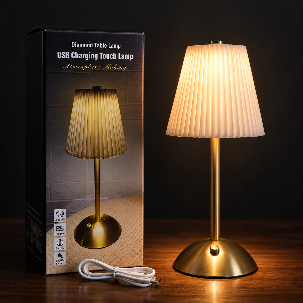 A table lamp with a pleated beige shade and a brushed brass base, shown lit next to its retail box and a white charging cable on a dark wooden surface against a black background.