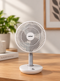 A small white rechargeable desktop fan with a silver stand, placed on a wooden table in a bright, modern room with a blurred plant and framed art in the background.