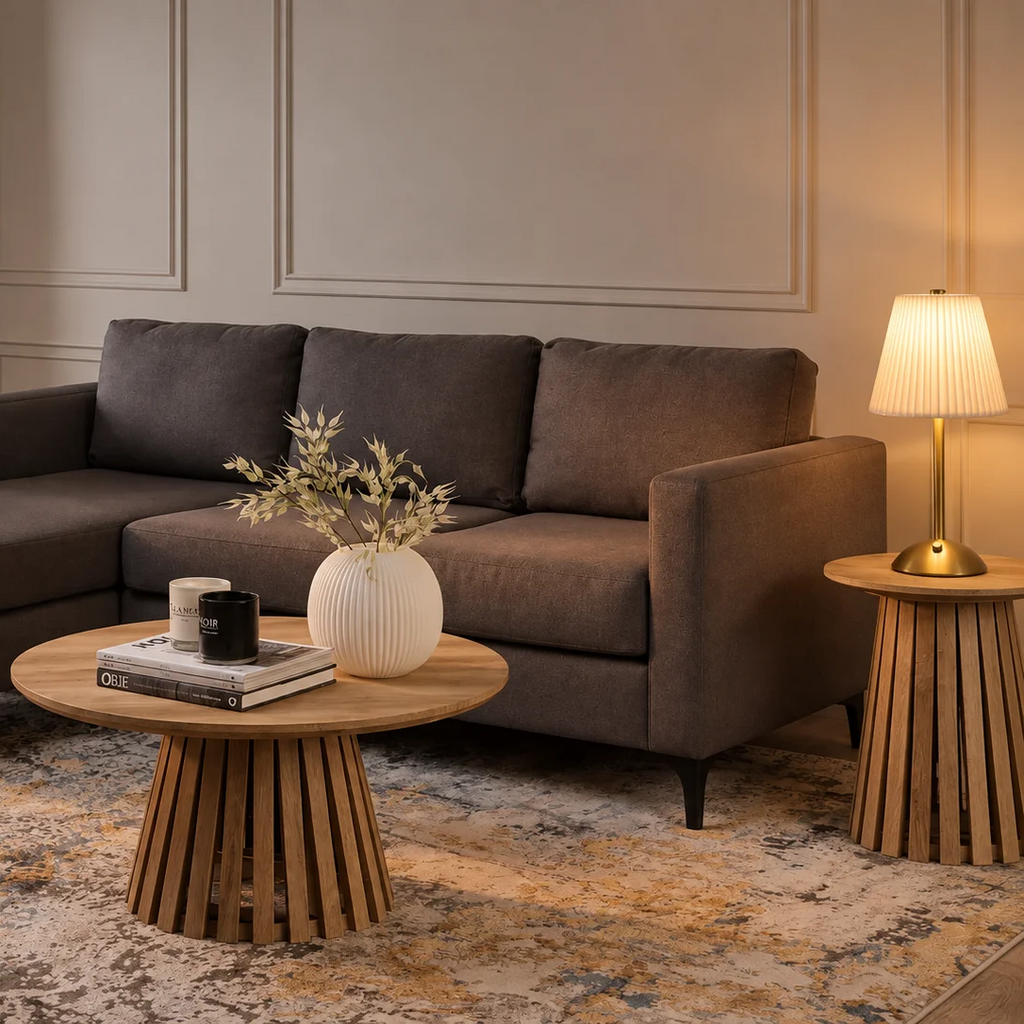 A pleated-shade table lamp glowing warmly on a side table next to a dark brown sofa in a styled living room, shown with the lamp fully in frame.
