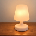A small modern table lamp with a frosted white shade and circular base, glowing warm yellow on a wooden surface against a neutral wall.