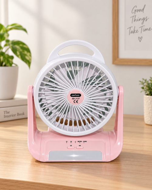 Pink and white portable desk fan with a handle, shown on a wooden tabletop in a bright indoor setting with plants and framed wall art in the background.