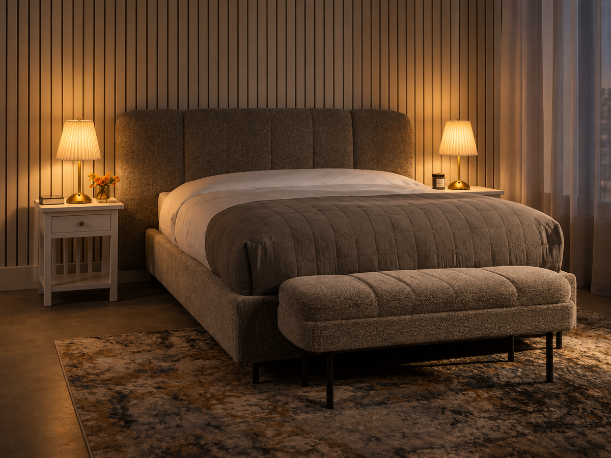 A modern upholstered bed in a warm-toned bedroom, featuring a tall segmented headboard, matching upholstered footboard bench, white bedding, two bedside tables with lit lamps, and a patterned rug.