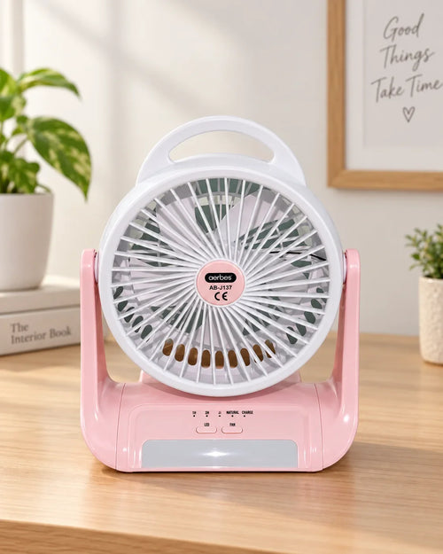 Pink and white portable desk fan with a handle, shown on a wooden tabletop in a bright indoor setting with plants and framed wall art in the background.